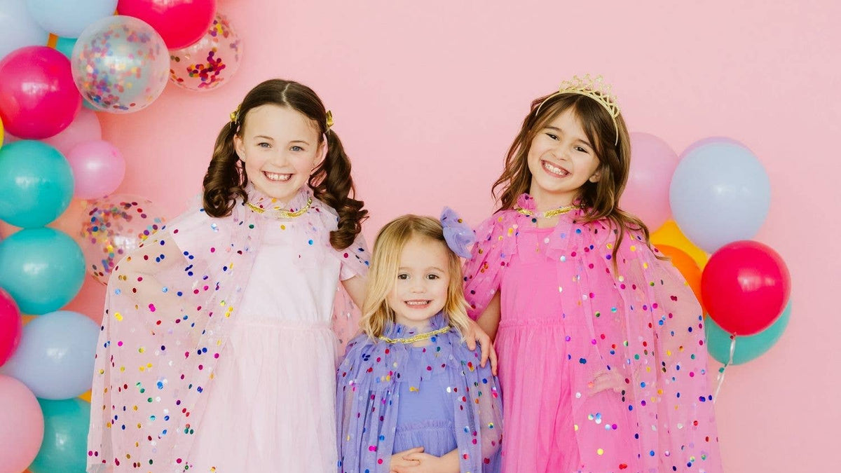 Three young girls in colorful dresses standing in front of balloons and confetti on a pink background