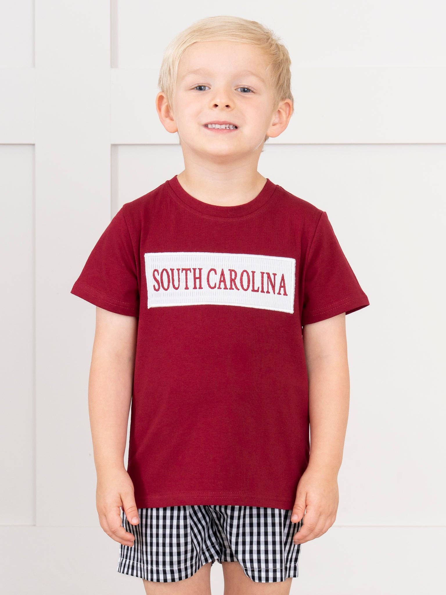 A young child wearing a maroon short-sleeve t-shirt with a white rectangular chest graphic that reads 'SOUTH CAROLINA' in maroon text, paired with black-and-white gingham shorts, standing against a white paneled wall.